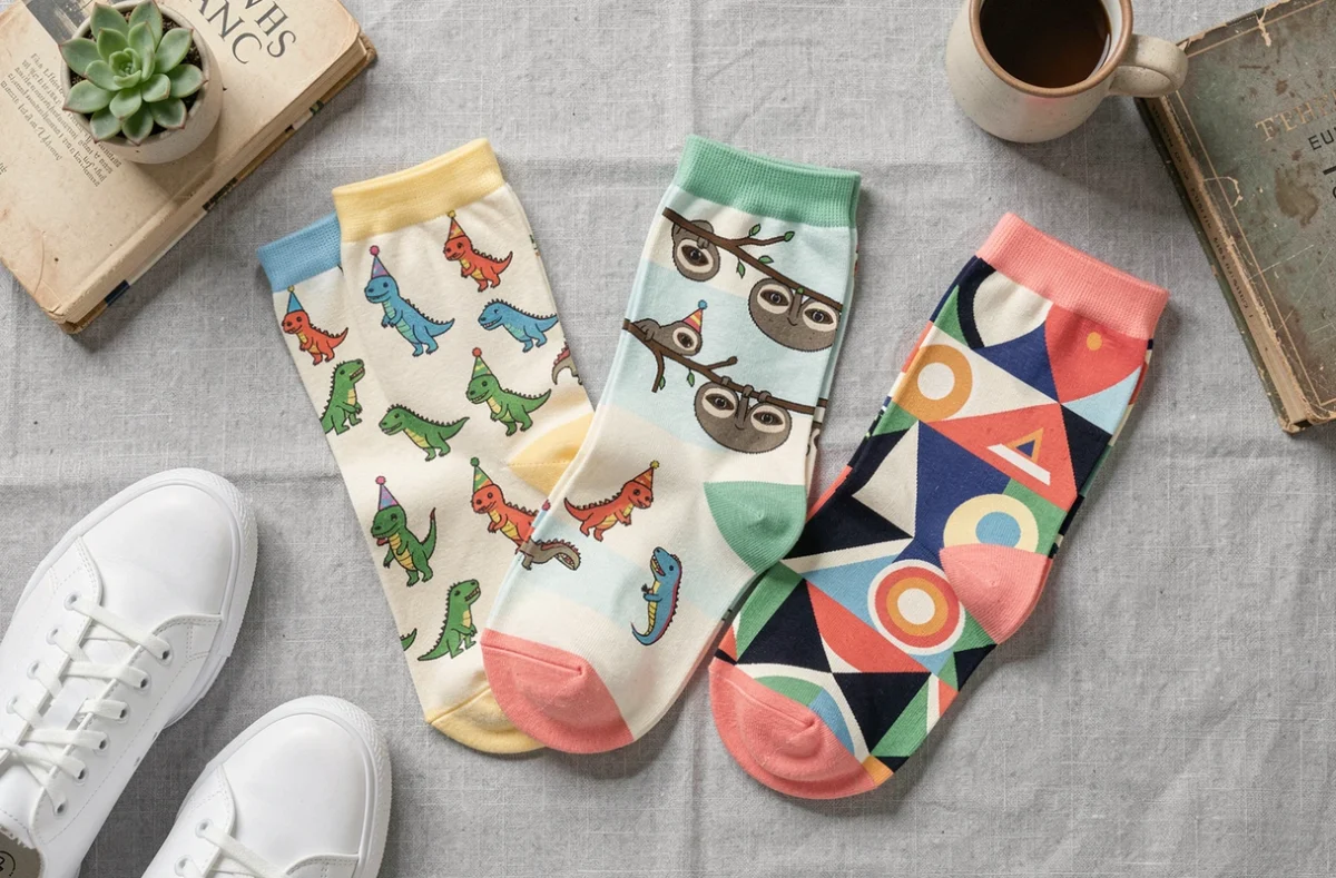 Flat-lay de chaussettes humoristiques femme avec motifs dinosaures, paresseux et géométriques retro, disposées sur fond lin gris avec sneakers et accessoires lifestyle